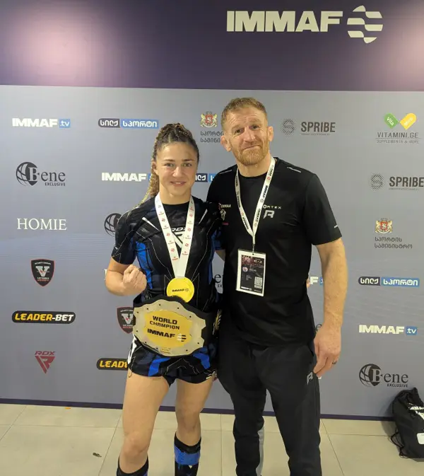 Katrina Fisher wearing her IMMAF World Champion belt and gold medal, standing with a coach
